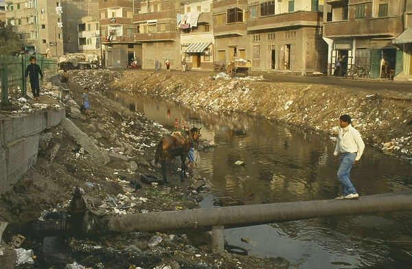 10048981. EGYPT Nile Delta Rubbish in canal with man walking over pipe