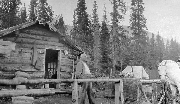 ALASKA: LOG CABIN. A man standing in front of a log cabin in Alaska. Photograph