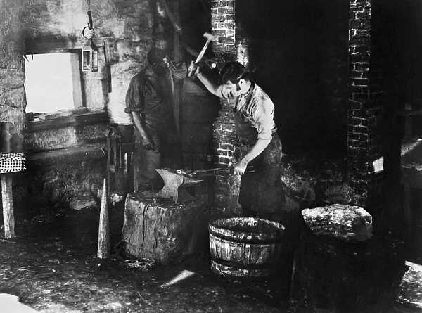 BLACKSMITH AT WORK. A blacksmith and assistant at work. Undated photograph