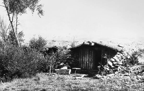 COLORADO: DUGOUT HOME. A frontiersman seated outside his dugout cabin in Grand County, Colorado