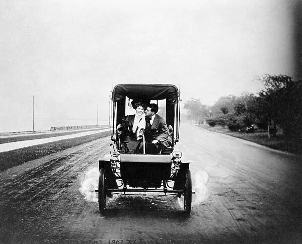 COUPLE DRIVING, c1907. A woman driving a car and talking with a man. Photograph, c1907
