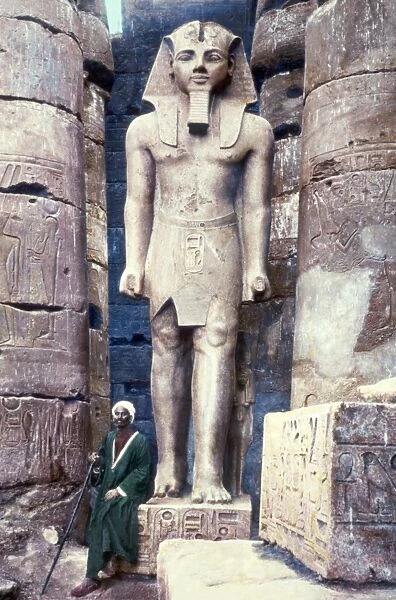 EGYPT: LUXOR TEMPLE. A man seated next to a statue of Pharaoh Ramesses II at the Luxor Temple