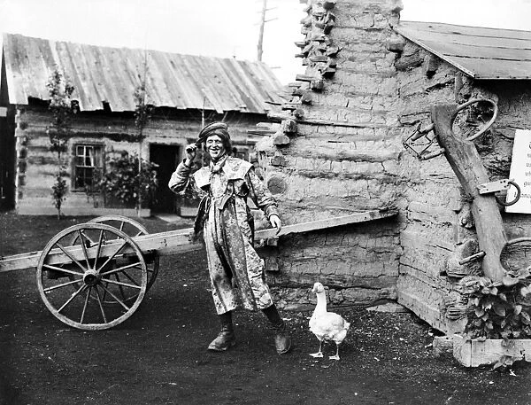 ENTERTAINER, 1901. An entertainer and a goose in a display of log cabins at the