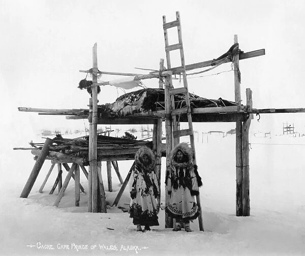 Two Eskimo women standing in front of a storage structure in Cape Prince of Wales, Alaska. Photographed by the Lomen Brothers, c1916