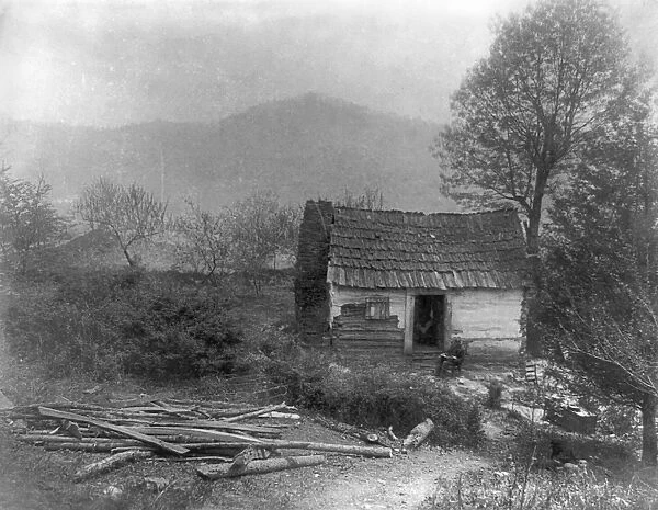 LOG CABIN, c1900. A man sitting in front of a small log cabin on a Sunday morning