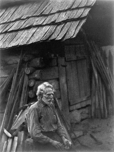 LOG CABIN, c1930. An old man sitting in front of a log cabin in rural America
