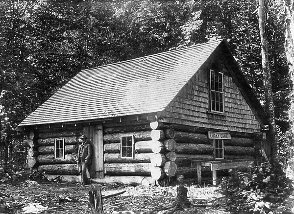 MAINE: LOG CABIN. A man standing in front of a log cabin at the Lucky Camp in Spencer Bay