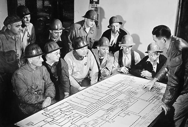 Men studying an oil flow chart showing the course of oil through a refinery, using the process of cracking, to supply fuel for American combat units fighting in World War II. Photograph, c1944