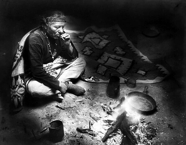 A Navajo man seated near a fire, smoking a cigarette after a meal. Photograph by William Carpenter, c1915