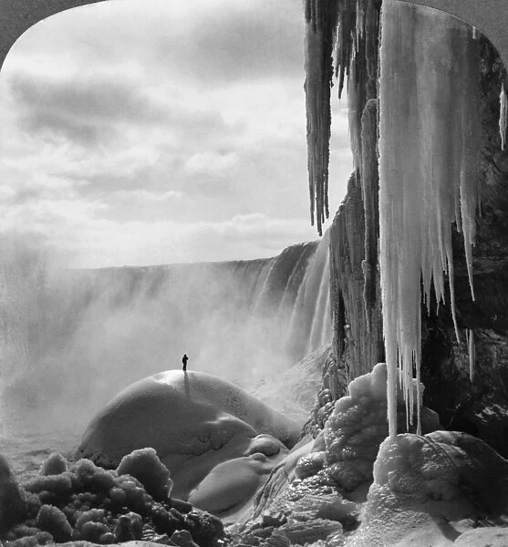 NIAGARA FALLS: FROZEN. Person standing on top of ice mound at Niagara Falls in winter, stereograph by R. Y. Young, c1903