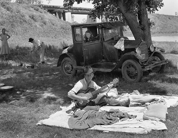 ROADSIDE CAMP, 1939. Migrant worker playing his guitar at a roadside camp for migrant workers