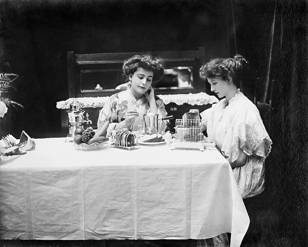 Two women seated at a breakfast table, using (left to right) an electric coffee pot, egg boiler, and toaster, all manufactured by General Electric. Photographed in 1908