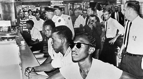 Four young black men sitting in at a segregated lunch counter at a drug store in Jackson, Mississippi, soon to be arrested by police officers (rear) for refusing an order to leave, 13 July 1961