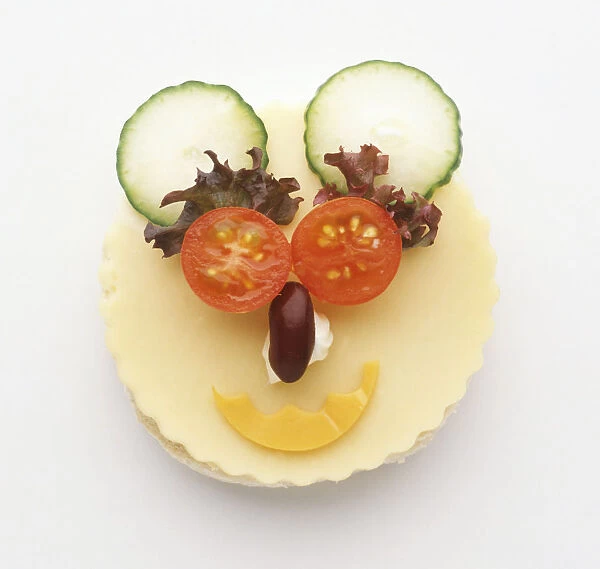 Face made from food using cheese covered muffin as a base, tomato eyes, lettuce eyebrows, kidney bean nose, cheese mouth, and cucumber ears