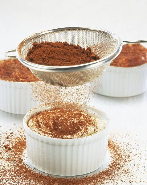 Grated chocolate being sprinkled from a sieve over a glass bowl containing Tiramisu