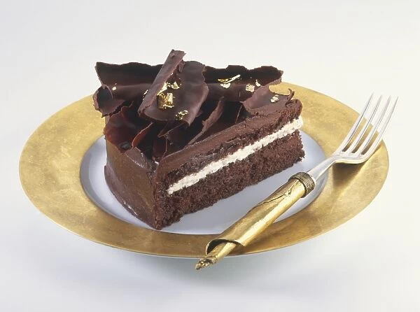 Slice of chocolate cake decorated with chocolate curls and pieces of gold leaf, served on gold plate with gold fork, side view