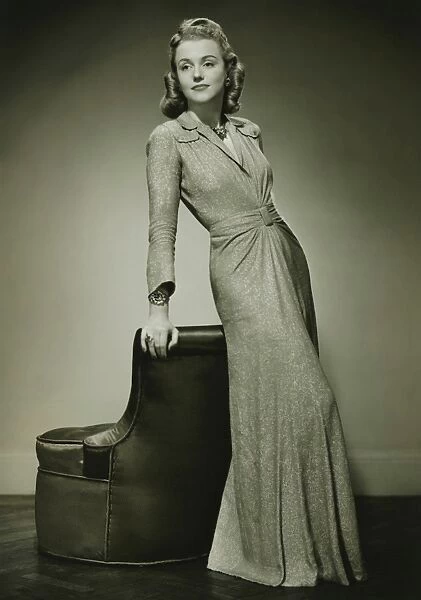 Young woman in evening dress leaning against chair, (B&W)