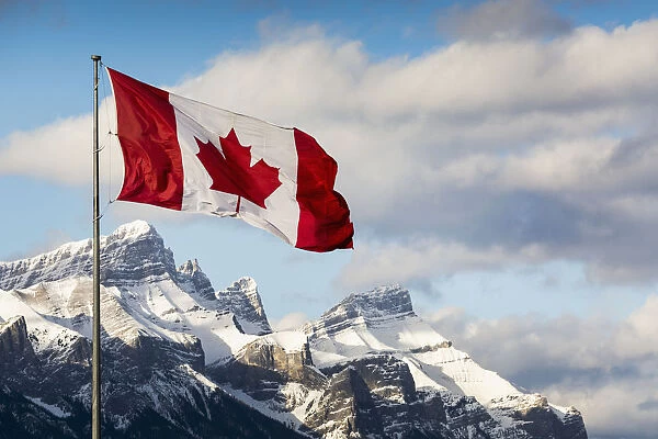 Canadian Flag Blowing In The Wind On A Flag Pole With Snow Covered Mountain Range In The Background With Blue Sky And Clouds; Canmore, Alberta, Canada