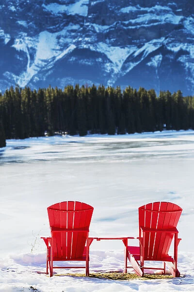 Two Red Chairs On Snow Covered Ridge Overlooking Frozen Lake With Snow Covered Mountain Cliff In The Background; Banff, Alberta, Canada