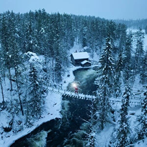 Santa Claus with lantern on the suspended bridge above the frozen rapids, Myllykoski, Juuma, Oulanka National Park, Kuusamo, Lapland, Finland (MR)