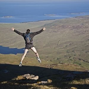 Fell runner jumping over rock (imitating base jump), coming down from mountain peak, with Loch an t-Siob in background