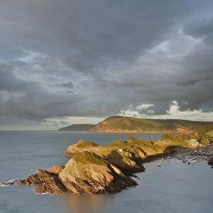 Rainclouds gathering over rocky coastal peninsula and bay in evening, with Great Hangmans and Combemartin in distance
