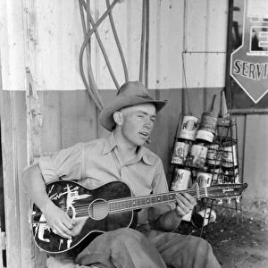 GUITAR PLAYER, 1940. A farm boy playing guitar outside of a filling station in Pie Town