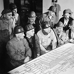 Men studying an oil flow chart showing the course of oil through a refinery, using the process of cracking, to supply fuel for American combat units fighting in World War II. Photograph, c1944