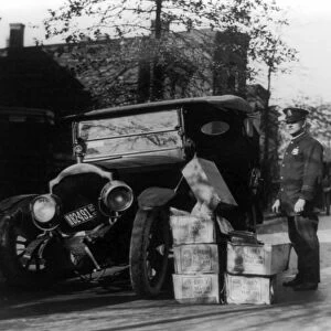 PROHIBITION, 1922. A police officer standing beside a wrecked car and cases of moonshine