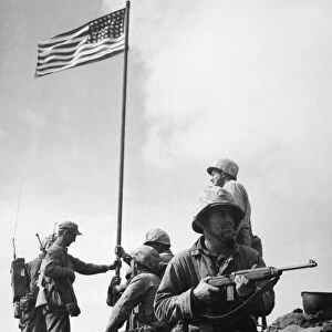 The raising of the first flag on Mount Suribachi, Iwo Jima, 23 February 1945. The soldiers are Sergeant Henry Hansen (without helmet), Sergeant Ernest Thomas (seated by flagpole), Corporal Charles Lindberg (standing at right), and Private First Class James Michels (on guard with carbine)