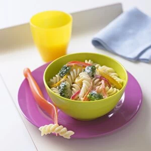 Bowl of fusilli pasta with broccoli, carrots, bell peppers in cheese sauce, fork, napkin and cup nearby