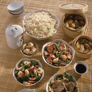 Bowls of Chinese food laid out on bamboo mat, including Dim sum, Szechuan lobster, stir-fried prawns and broccoli, pork stir-fry, Szechuan aubergine, rice, fortune cookies and tea pot