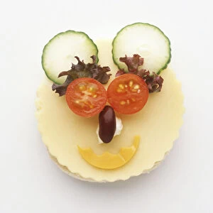 Face made from food using cheese covered muffin as a base, tomato eyes, lettuce eyebrows, kidney bean nose, cheese mouth, and cucumber ears