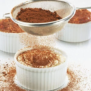 Grated chocolate being sprinkled from a sieve over a glass bowl containing Tiramisu