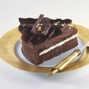 Slice of chocolate cake decorated with chocolate curls and pieces of gold leaf, served on gold plate with gold fork, side view