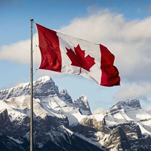 Canadian Flag Blowing In The Wind On A Flag Pole With Snow Covered Mountain Range In The Background With Blue Sky And Clouds; Canmore, Alberta, Canada