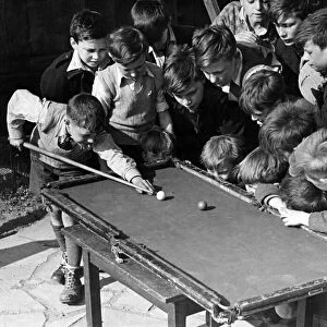 Children gather around a snooker table to play. Sixteen tousled heads bend over a rickety