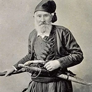 Three-quarter length portrait of an elderly greek. He is wearing traditional attire and has a pistol and a large dagger in a sheath. He is wearing a soft hat